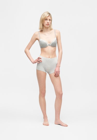 Kvinde stående barfodet iført en lys grå bh med blonder og matchende højtaljede bomuldsshorts på ensartet hvid baggrund.