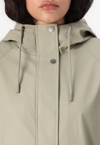 Veste imperméable vert clair avec une fermeture éclair et des boutons pression, dotée d'une capuche, d'un cordon de serrage et d'accents en métal. Texture lisse.