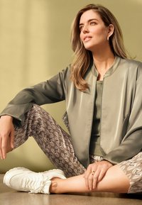 Giacca bomber in satin verde chiaro sopra un top aderente oliva, abbinata a pantaloni corti grigi con motivo e sneakers bianche. Anello d'oro sulla mano.