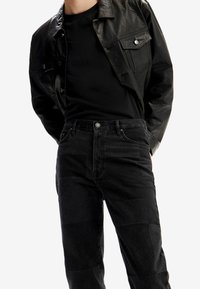 Giacca di pelle nera sopra una maglietta nera, abbinata a jeans neri con un design patchwork e chiusura con bottone in vita.