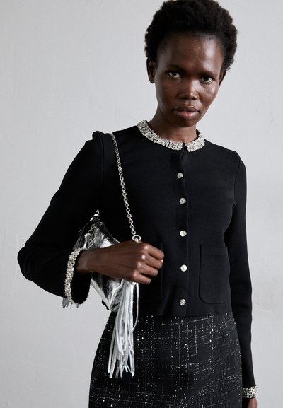 Svart figursydd jacka med långa ärmar, dekorerad krage, silverknappar och framfickor. Matchad med en strukturerad svart kjol och en metallisk handväska.