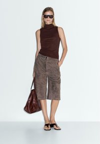Top sans manches en turtleneck marron, shorts marron en daim jusqu'aux genoux, tongs noires, et un grand sac à main en cuir marron. Textures lisses.