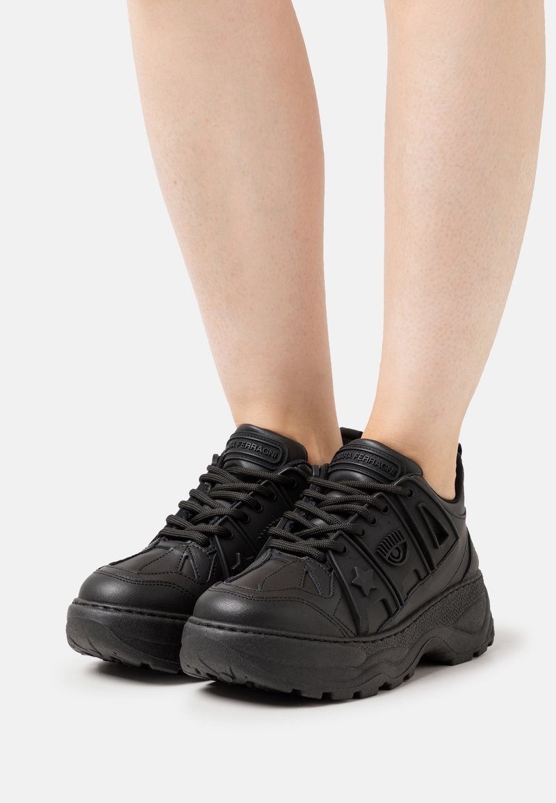 Svarta syntetsneakers med texturerade detaljer, snördesign, kraftig sula och logotypdetaljer på tungan och sidopanelerna.