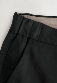 Schwarze Hose mit glatter Textur und flachem Taillenbund. Sichtbare Nähte entlang des Taillenbundes und der Seitennaht.