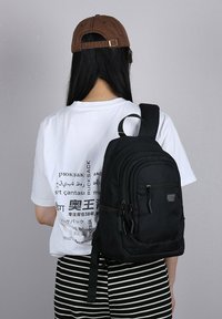 Zaino in tessuto nero con più scomparti con zip, tracolla regolabile e maniglia superiore. Abbinato a una t-shirt bianca con grafica e pantaloni a righe.