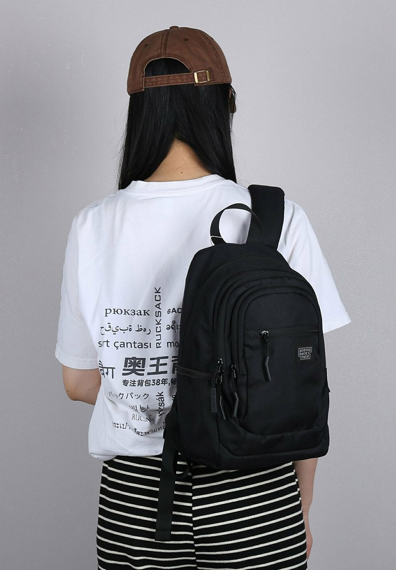 Zaino in tessuto nero con più scomparti con zip, tracolla regolabile e maniglia superiore. Abbinato a una t-shirt bianca con grafica e pantaloni a righe.