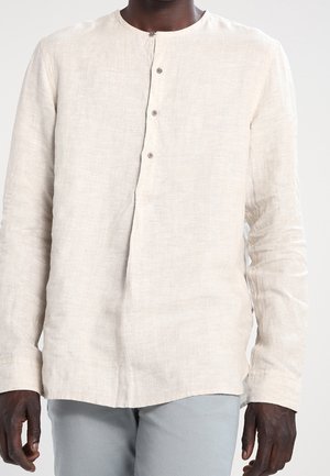 Hombre vistiendo una camisa de lino de manga larga, color beige claro, abotonada, combinada con pantalones gris claro, sobre un fondo liso.