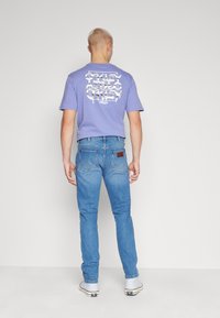 Lila t-shirt med vit grafisk design på ryggen, tillsammans med ljusblå jeans. Modellen bär vita sneakers. Enkel, avslappnad outfit.