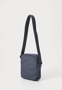 Námořnická modrá crossbody taška vyrobená z hladké tkaniny, s nastavitelným popruhem a značkováním "TOMMY JEANS" a zipovým uzávěrem.
