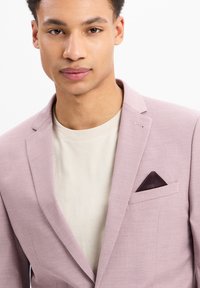 Helles pinkes Blazer mit Reverskragen, einer einzelnen Brusttasche, in der ein dunkelviolettes Taschentuch steckt, getragen über einem beigen Rundhals-T-Shirt.