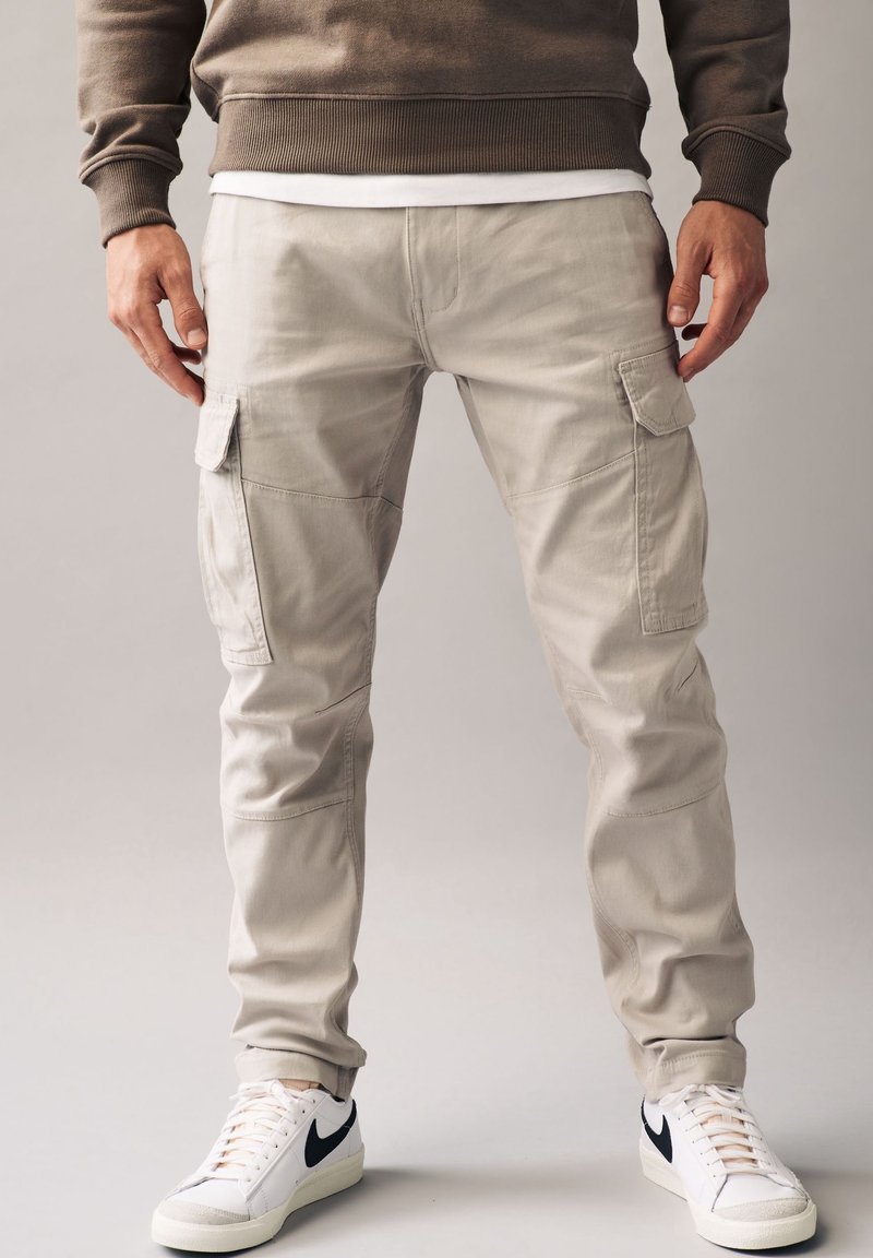 Homme portant un pantalon cargo beige et des baskets blanches avec un logo Nike noir, debout devant un fond clair uni.