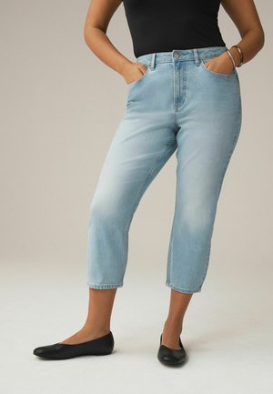 Kobieta ubrana w jasnoniebieskie krótkie dżinsy, czarny bezrękawnik, czarne baleriny i srebrne bransoletki, z rękami w kieszeniach, stojąca na neutralnym tle.