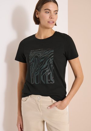 Frau trägt ein schwarzes T-Shirt mit kurzen Ärmeln, abstraktem Tierprint und Text, kombiniert mit beigen Hosen, steht mit einer Hand in der Tasche.