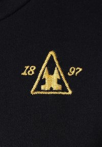 Tela negra con bordado dorado que presenta un diseño triangular, números '1897' y botas estilizadas en el centro.