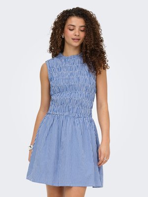 Vrouw met krullend haar die een mouwloze blauw-witte geruite jurk draagt met een geplooide lijfje en een uitlopende rok, staand tegen een effen achtergrond.