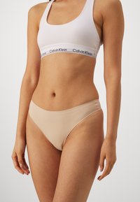 Beige hipster-alushousut sileällä kankaalla, matalalla vyötäröllä ja vähillä saumoilla. Yhdistetty valkoiseen rintaliiviin, jossa on musta logonauha.