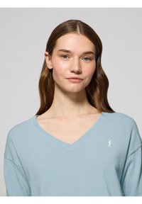 Maglione azzurro chiaro a V in tessuto morbido con vestibilità rilassata. Presenta un piccolo logo ricamato sul petto e un colletto a costine.