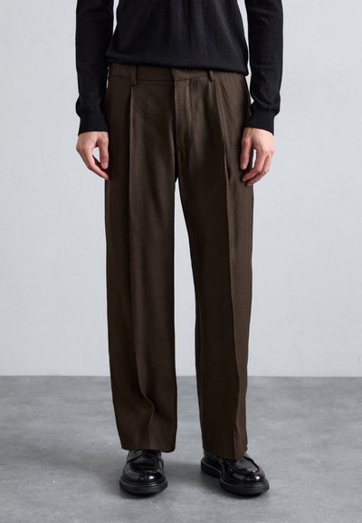 Pantaloni marroni plissettati in tessuto strutturato, con una vestibilità comoda e passanti per cintura, abbinati a scarpe nere lucide.