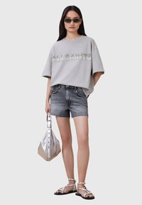 Grå oversized T-shirt med korte ærmer, hvor der står "ALLSAINTS" i rhinsten, parret med mørk denim shorts, sølv taske og flade sandaler.