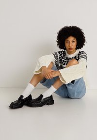 Paksu kreemja kabelikootud kampsun mustade geomeetriliste mustritega, laiad sinised teksaseelikud, valged sokid ja mustad loaferid, istub valgel pinnal.