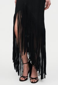 Μαύρη φούστα με ασύμμετρη λεπτομέρεια με κρόσσια που αποκαλύπτουν τα πόδια, σε συνδυασμό με μαύρες ψηλοτάκουνες σανδάλες με λουράκια, σε φωτεινό φόντο.