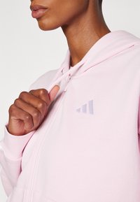 Světle růžová zipová mikina vyrobená z tkaniny se směsí bavlny. Má vyšívané logo se třemi naskládanými pruhy v levé části hrudi.