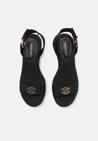 Black suede heeled sandals with a closed back, adjustable ankle strap, and a decorative gold-tone metal accent on the front strap.