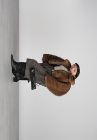 Person wearing a brown fur coat, gray wide-leg pants, black boots, holding a black handbag, standing against a plain light background.