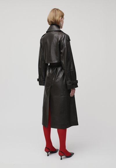 Frau in einem schwarzen Leder-Trenchcoat, roten Strumpfhosen, schwarzen spitzen High Heels und großen silbernen Ohrringen, die vor einem neutralen Hintergrund steht.