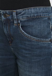 Jeans de mezclilla azul oscuro con costuras naranjas, que cuentan con un bolsillo delantero y un cierre de botón. La tela tiene una textura suave con un ligero desgaste.