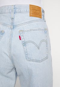 Jeans de mezclilla azul claro con una etiqueta de cuero en la cintura y una pestaña roja en el bolsillo trasero, con costuras en contraste y un diseño curvado.
