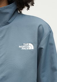 Κοντινό πλάνο ενός ατόμου που φορά ένα μπλε μπουφάν με ψηλό γιακά και το λευκό λογότυπο The North Face στο στήθος.