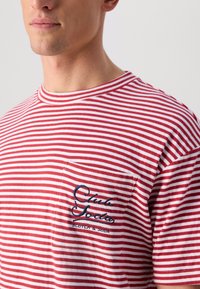 Rød-og-hvitstripet t-skjorte med en lomme som har blå broderte tekst "Club Soda Scotch & Soda," laget av mykt bomullsmateriale.