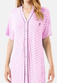 Camisón de color rosa claro con mangas cortas, frente abotonado, pequeño patrón de corazones y un bolsillo en el pecho que presenta un logo. Textura de tejido suave.