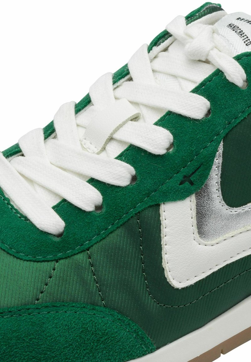 Grüne Turnschuhe aus einer Mischung aus Wildleder und glattem Stoff, mit weißen Schnürsenkeln und einem silbernen Akzent nahe der Ferse. Sichtbare gestickte Details.