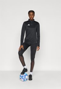 Μαύρο αθλητικό τοπ με φερμουάρ στον γιακά και λευκό λογότυπο, σε συνδυασμό με μαύρο κολάν που έχει διαφανείς λεπτομέρειες και λευκές ρίγες, στεκόμενη πάνω σε μια μπάλα ποδοσφαίρου.