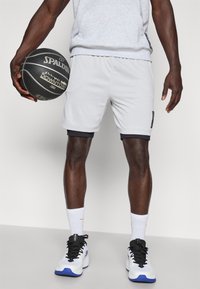 Šedé športové šortky s čiernym lemom a bočným logom, spárované s bielymi ponožkami a čierno-bielymi teniskami, drží čiernu basketbalovú loptu.
