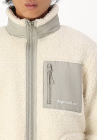 Hvid fleecejakke med grå detaljer, høj krave, frontlynlås og brystlomme med et logo. Struktureret, warm og hyggeligt materiale.