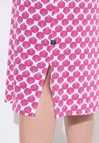 Tessuto con motivo a conchiglia rosa presenta una fessura laterale, evidenziando una superficie liscia e uno sfondo bianco. È visibile un piccolo etichetta nera.