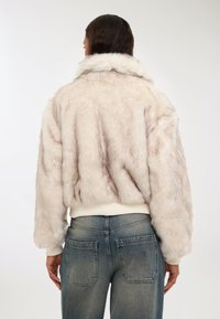 Giacca in pelliccia sintetica di un beige chiaro con texture morbida, collo ampio e polsini a coste. Abbinata a jeans a vita alta in blu sbiadito.