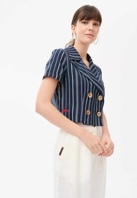 Granatowa krótka kurtka z białymi pionowymi paskami, podwójnym zapięciem, drewnianymi guzikami i krótkimi rękawami. Czerwony akcent po bokach.