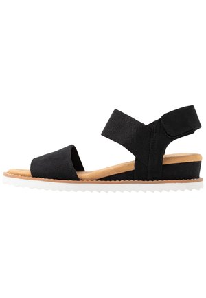 Sorte sandaler med tekstilremme, en blød tan-fodbed og en hvid, struktureret ydersål. Har to brede justerbare remme for komfort.