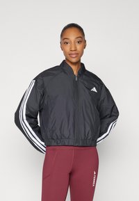 Črna bomberska jakna z zadrgo spredaj, elastičnim robom in belim dizajnom s tremi črtami na rokavih. Material se zdi gladek in lahek.