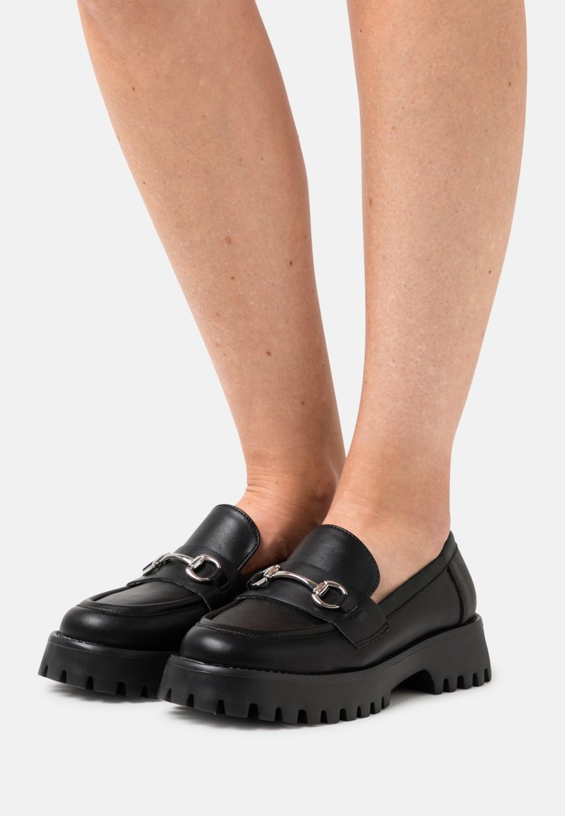Mocassins en cuir noir à semelles épaisses avec détail en métal argenté, portés pieds nus sur un fond gris clair uni.