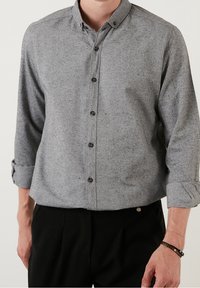 Grijs overhemd met knopen van een gestructureerde stof, voorzien van een brede kraag, lange mouwen met opgerolde manchetten en donkere knopen.