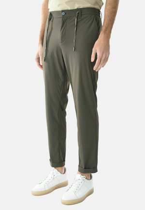 Uomo che indossa pantaloni aderenti verde oliva con risvolti arrotolati e sneaker bianche, in piedi davanti a uno sfondo semplice.