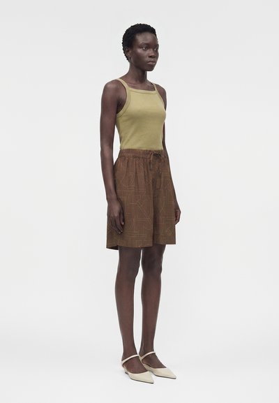 Μαύρη γυναίκα που στέκεται φορώντας πράσινο αμάνικο τοπ, καφέ φούστα με σχέδιο και κορδόνια, και μπεζ μυτερές μπαλαρίνες σε λευκό φόντο.