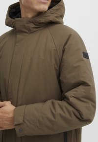 Smeđa, izolirana jakna s visokim ovratnikom i podesivom kapuljačom. Ima bočne patent-zatvarače i teksturiranu tkaninu bez vidljivih uzoraka.
