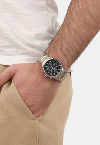 Silberner Metall-Zeitmesser mit schwarzem Zifferblatt, Chronographenfunktionen und einem Armband aus Edelstahl, getragen am Handgelenk einer Person in hellbeigen Hosen.