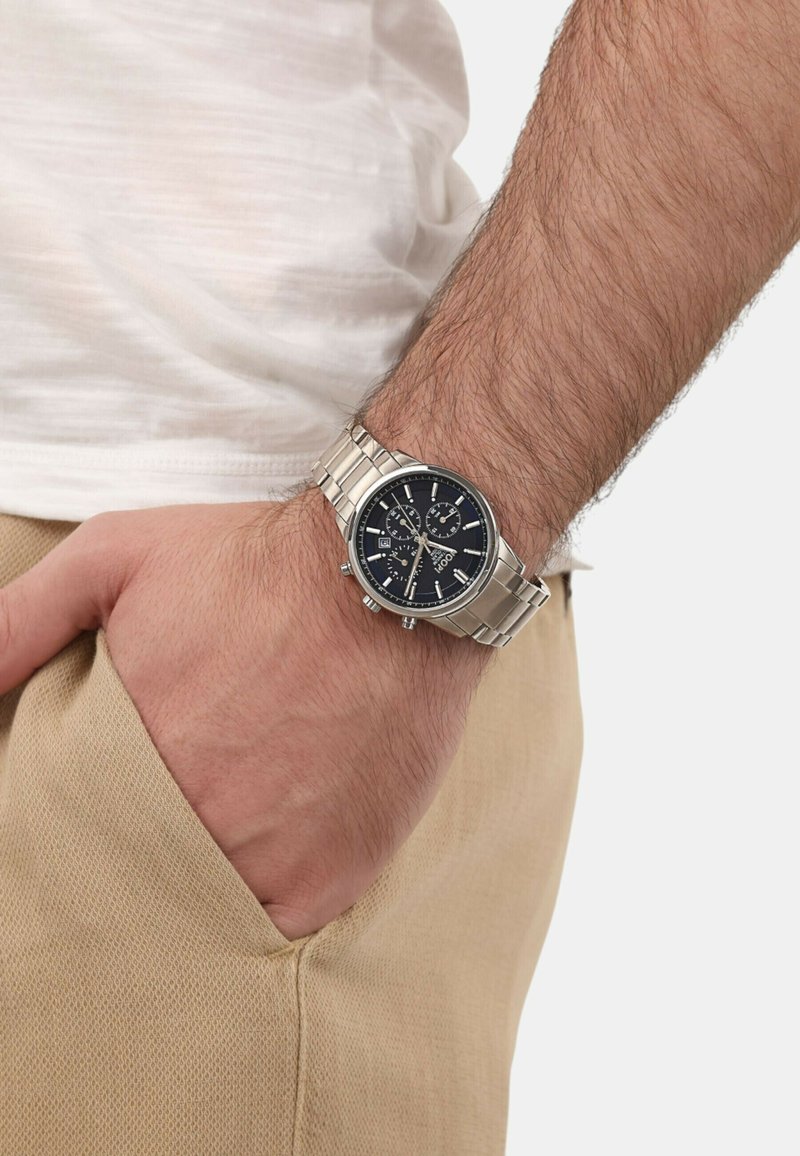 Silberner Metall-Zeitmesser mit schwarzem Zifferblatt, Chronographenfunktionen und einem Armband aus Edelstahl, getragen am Handgelenk einer Person in hellbeigen Hosen.